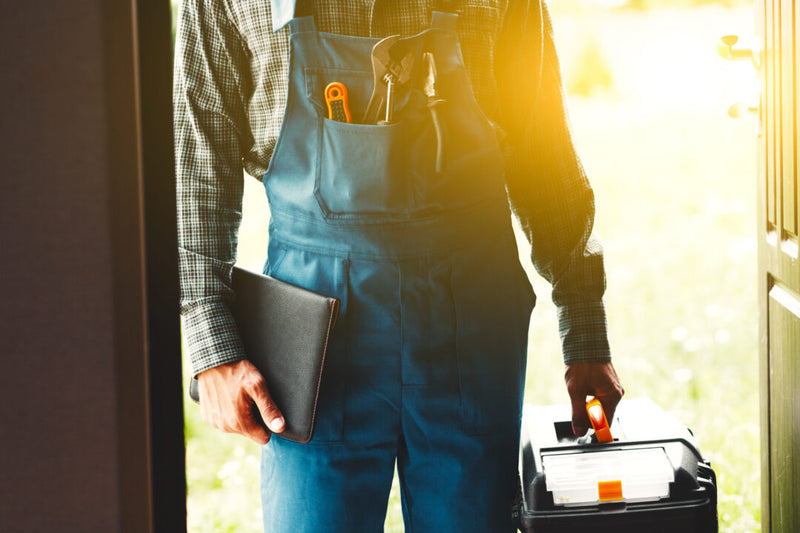 Person wearing blue overalls holding a toolbox and a notebook outdoors.